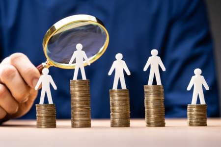 Person using magnifying glass to examine pay disparity, represented by stacks of coins